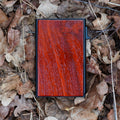 Nahaufnahme eines flachen, rechteckigen Kartenetuis mit einer Frontplatte aus intensiv rot gemasertem Holz, eingebettet in trockenes, braunes Eichenlaub.
