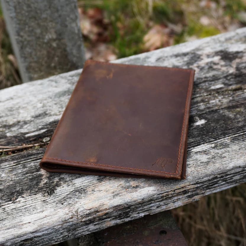 Ein schlichtes, flaches Portemonnaie aus dunkelbraunem Leder mit dezenter Logo-Prägung, liegend auf einer verwitterten Holzbank im Freien.