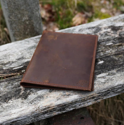 Ein schlichtes, flaches Portemonnaie aus dunkelbraunem Leder mit dezenter Logo-Prägung, liegend auf einer verwitterten Holzbank im Freien.