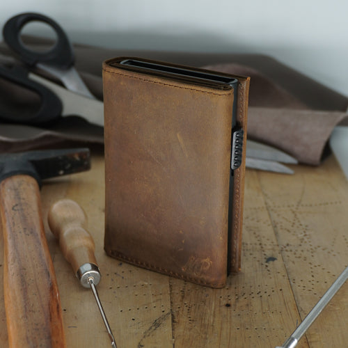 Braunes Vintage-Leder-Portemonnaie mit Metall-Kartenhalter auf einem Holzwerktisch, umgeben von Schusterwerkzeugen wie Hammer, Scheren und Lederresten, handgefertigte Qualität symbolisierend.