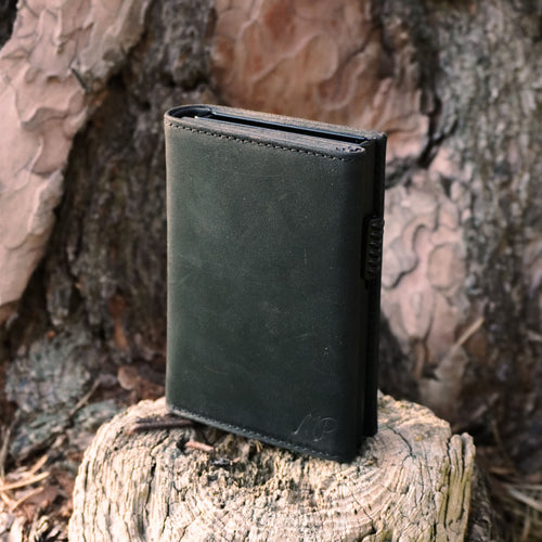 Ein elegantes, dunkelgraues oder schwarzes Leder-Wallet, aufrecht stehend auf dem hellen Anschnitt eines Baumstammes vor rissiger Rinde.