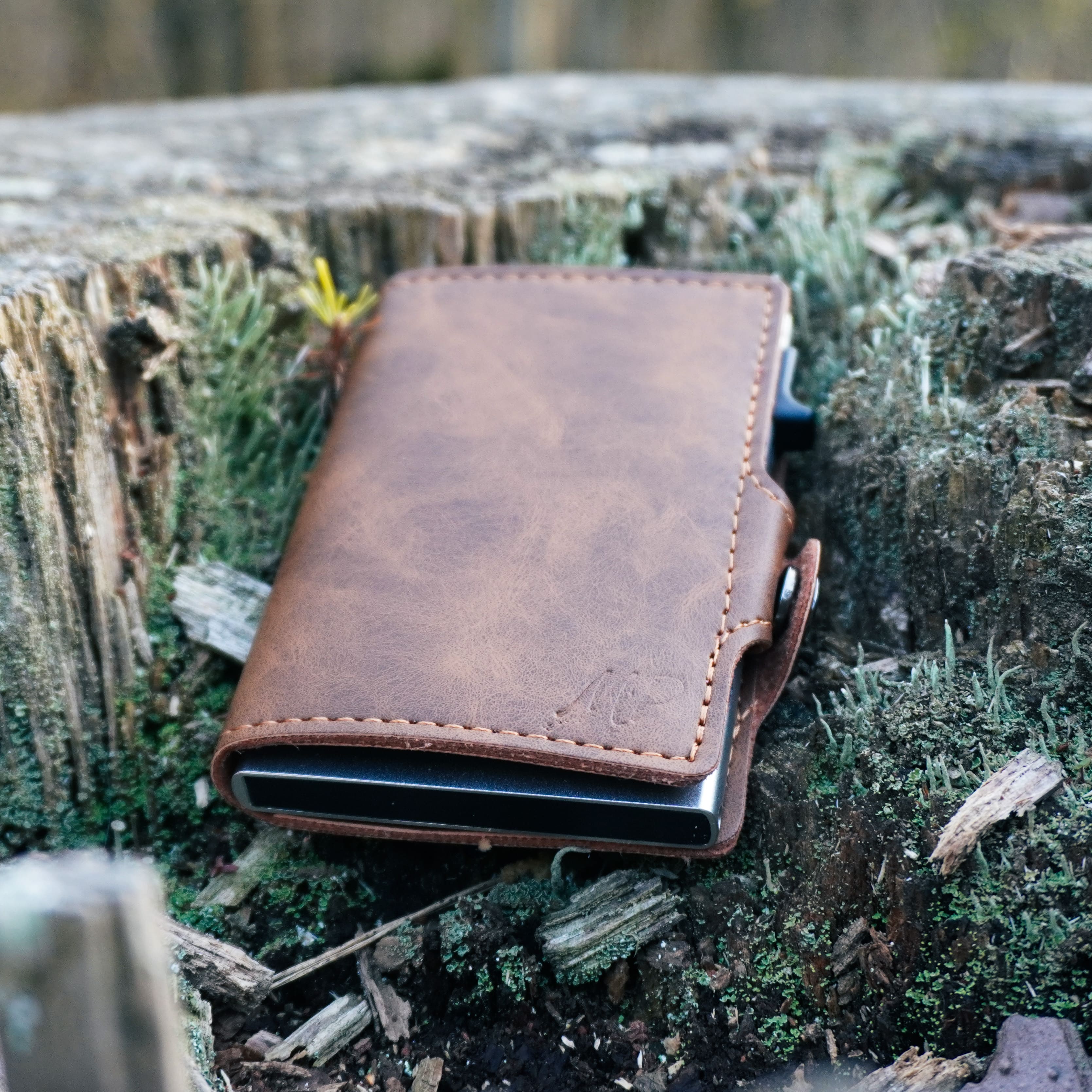 Ein kompaktes, hellbraunes Leder-Wallet mit sichtbarer Naht und seitlichem Schiebemechanismus, platziert auf einem moosigen Baumstumpf im Wald.